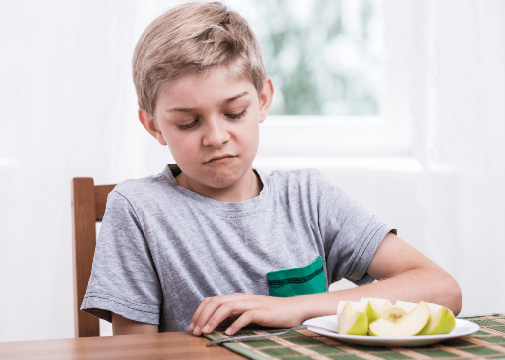 Child sitting at a table with left arm resting on table looking disgusted at a plate of apple slice. 