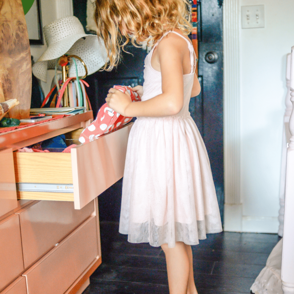 Girl in a pink dress standing at dresser looking at clothes in the top drawer. 